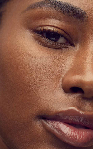 A close-up photo of a Black woman with smooth, glowing skin. She appears to have just moisturized her face, as her skin looks hydrated and radiant. The photo is well-lit and the woman is surrounded by a blurred background.