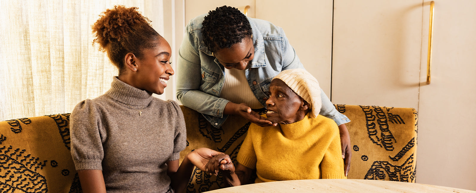 Two younger women engaging warmly with an older woman seated on a sofa, one leaning in close to her face