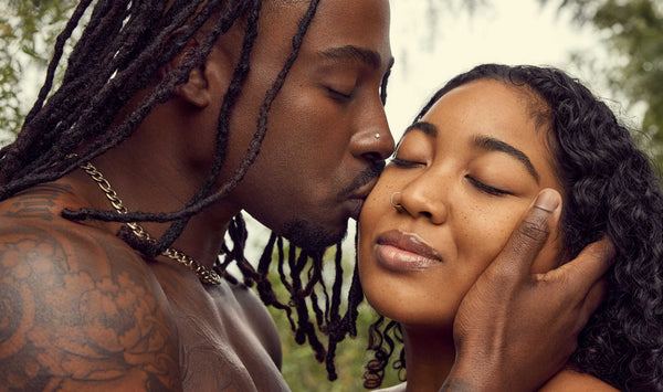 An outdoor photo of a Black couple. The man is kissing the woman on the cheek, with their eyes closed in a moment of affection. They are standing close to each other and holding hands, with a beautiful natural background of trees and sunlight behind them.  Both look happy and relaxed, enjoying the moment. The photo is well-lit and captures the warmth and tenderness of the scene.